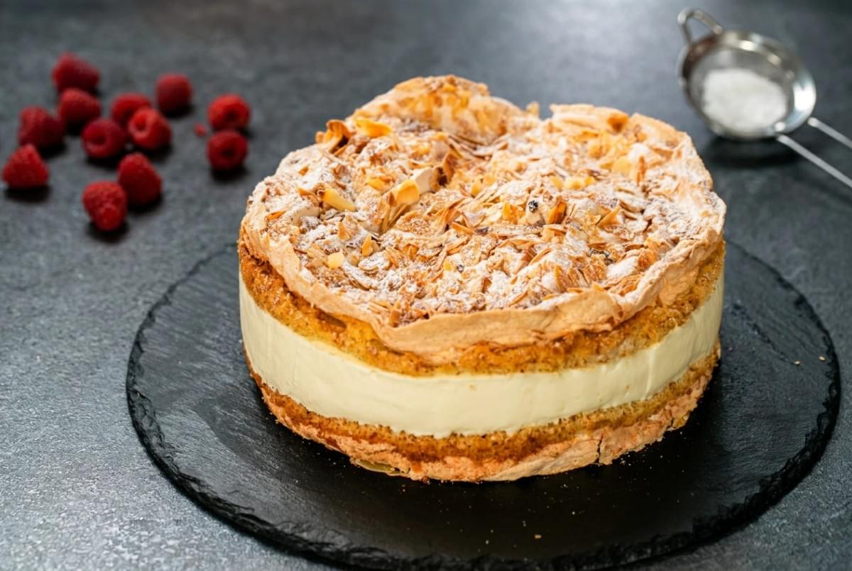 Round meringue layer cake topped with toasted sliced almonds, dusting sugar, and fresh raspberries on a slate tray. Round meringue layer cake topped with toasted sliced almonds, dusting sugar, and fresh raspberries on a slate tray.