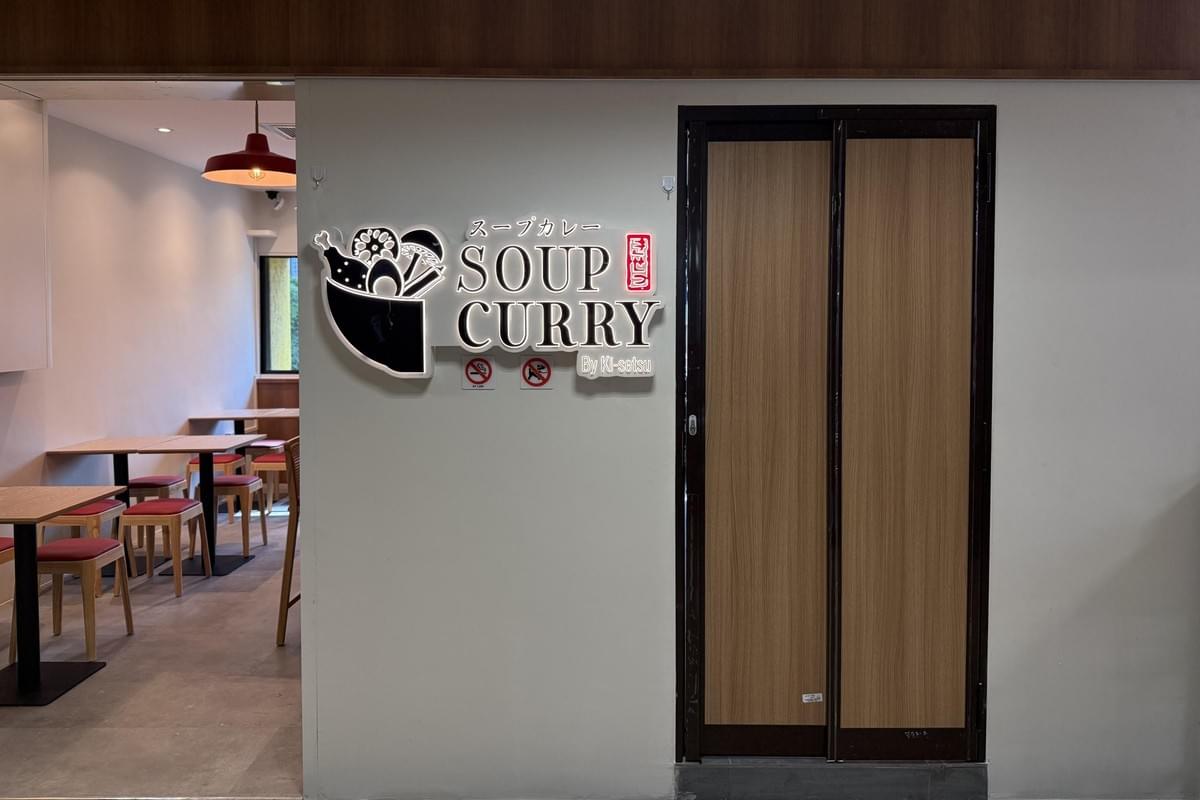A illuminated wall sign displaying "Soup Curry By Ki-setsu" and a bowl illustration sits next to a closed wooden sliding door. To the left, an open doorway reveals a brightly lit restaurant seating area with wooden tables and red-cushioned chairs. A illuminated wall sign displaying "Soup Curry By Ki-setsu" and a bowl illustration sits next to a closed wooden sliding door. To the left, an open doorway reveals a brightly lit restaurant seating area with wooden tables and red-cushioned chairs.