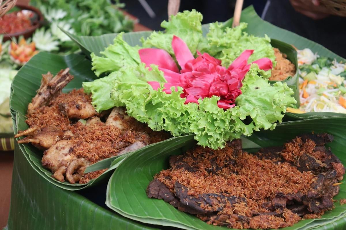 A large platter lined with fresh green banana leaves features fried chicken pieces topped with toasted coconut serundeng alongside other dishes. Bright green lettuce and a carved red torch ginger flower garnish center the vibrant Indonesian food arrangement. A large platter lined with fresh green banana leaves features fried chicken pieces topped with toasted coconut serundeng alongside other dishes. Bright green lettuce and a carved red torch ginger flower garnish center the vibrant Indonesian food arrangement.