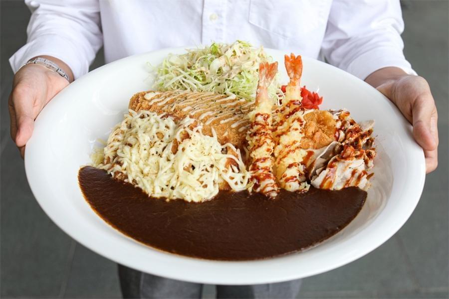 A person holds an enormous white plate piled high with a massive serving of Japanese curry pool-layered with a rich, dark brown sauce. The dish is loaded with an array of toppings, including melted cheese over rice, fried cutlets, two crispy tempura shrimp, sliced pork, and a mountain of shredded cabbage. A person holds an enormous white plate piled high with a massive serving of Japanese curry pool-layered with a rich, dark brown sauce. The dish is loaded with an array of toppings, including melted cheese over rice, fried cutlets, two crispy tempura shrimp, sliced pork, and a mountain of shredded cabbage.