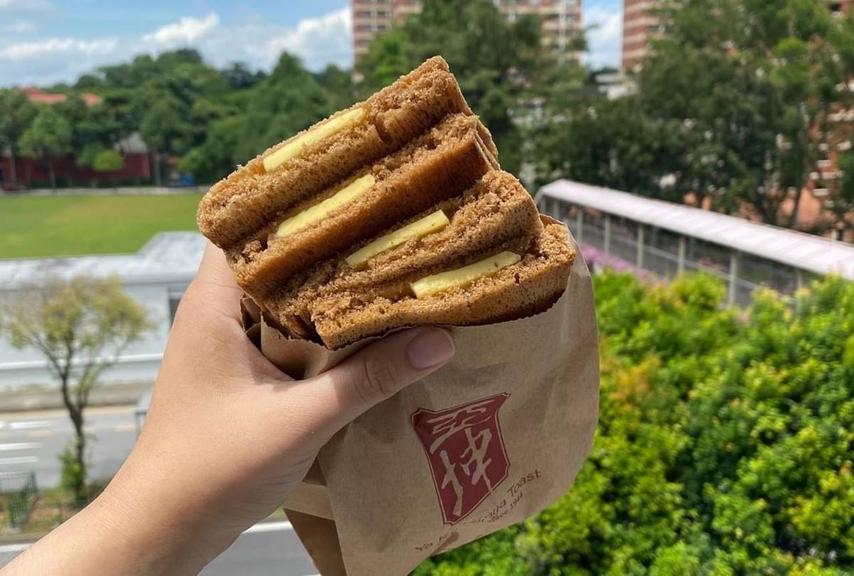 This image shows a person holding a brown paper bag filled with several slices of toasted Kaya bread. Each slice of dark, crispy toast is layered with visible pats of yellow butter, held up against an outdoor background of lush green trees and buildings. This image shows a person holding a brown paper bag filled with several slices of toasted Kaya bread. Each slice of dark, crispy toast is layered with visible pats of yellow butter, held up against an outdoor background of lush green trees and buildings.