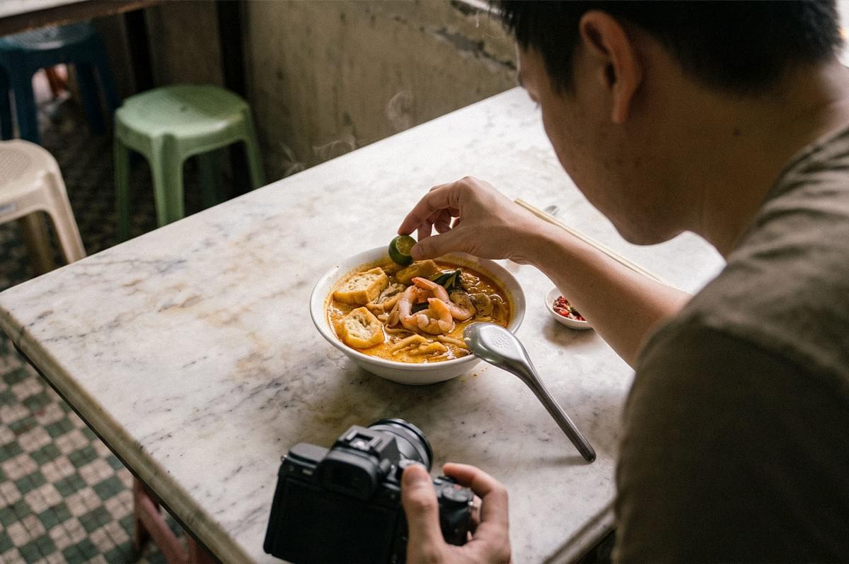 A man photographs a bowl of noodle soup while adding a lime wedge to the dish, with his camera resting on the table. A man photographs a bowl of noodle soup while adding a lime wedge to the dish, with his camera resting on the table.