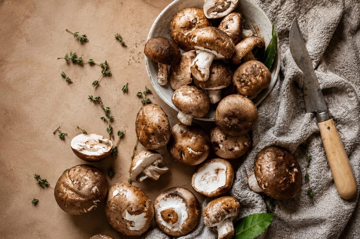Fresh cremini mushrooms scattered on a neutral surface with a ceramic bowl, paring knife, and fresh thyme sprigs. Fresh cremini mushrooms scattered on a neutral surface with a ceramic bowl, paring knife, and fresh thyme sprigs.