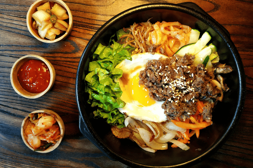 A top-down view of bibimbap served in a black stone bowl (dolsot). The dish includes prominent mounds of bulgogi, lettuce, and onions, served alongside three small side dishes (banchan) containing kimchi, pickled onions, and a small bowl of red gochujang sauce. A top-down view of bibimbap served in a black stone bowl (dolsot). The dish includes prominent mounds of bulgogi, lettuce, and onions, served alongside three small side dishes (banchan) containing kimchi, pickled onions, and a small bowl of red gochujang sauce.