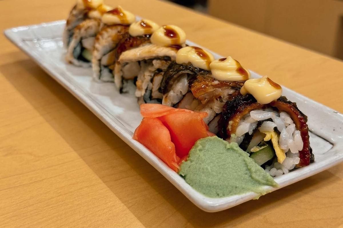 This close-up shot features an unagi sushi roll lined up diagonally across a long, white rectangular ceramic plate set on a light wooden table. The roll is stuffed with cucumber and tamago, draped in glazed grilled eel, and finished with creamy dollops of mayonnaise and sweet unagi sauce on each piece, accompanied by pink pickled ginger and a smear of green wasabi paste in the foreground. This close-up shot features an unagi sushi roll lined up diagonally across a long, white rectangular ceramic plate set on a light wooden table. The roll is stuffed with cucumber and tamago, draped in glazed grilled eel, and finished with creamy dollops of mayonnaise and sweet unagi sauce on each piece, accompanied by pink pickled ginger and a smear of green wasabi paste in the foreground.