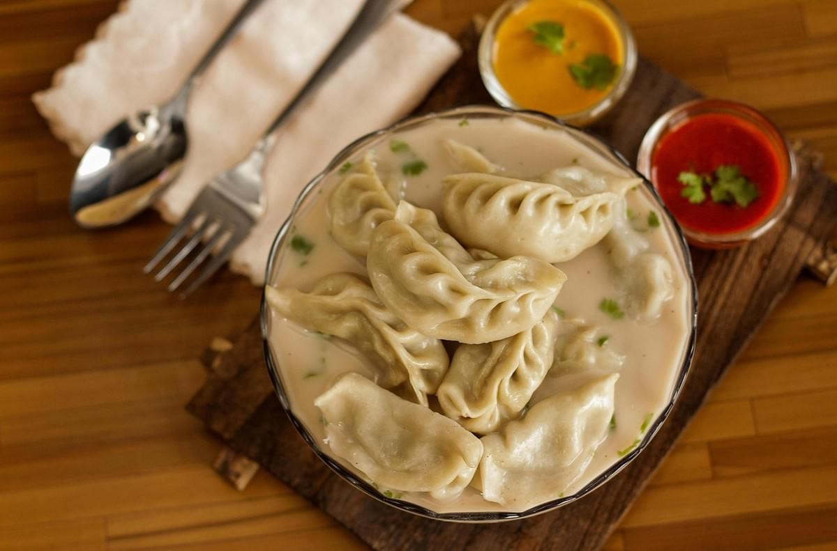 A large clear bowl is filled with pleated dumplings submerged in a creamy, light-colored broth and garnished with fresh cilantro. The bowl sits on a rustic wooden board alongside a fork, spoon, and two small dishes containing vibrant red and yellow dipping sauces. A large clear bowl is filled with pleated dumplings submerged in a creamy, light-colored broth and garnished with fresh cilantro. The bowl sits on a rustic wooden board alongside a fork, spoon, and two small dishes containing vibrant red and yellow dipping sauces.