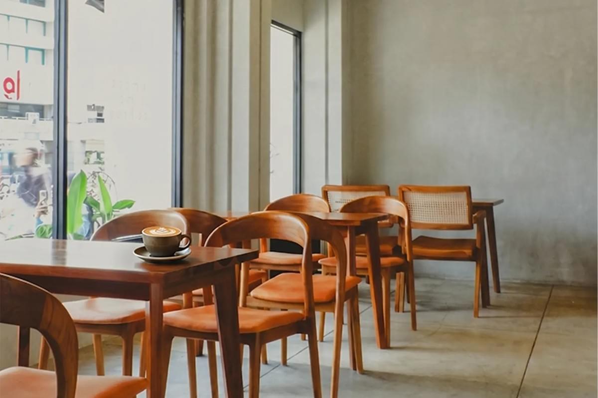 Minimalist cafe interior featuring wooden tables and chairs, a concrete wall, and a cup of latte art by a large window. Minimalist cafe interior featuring wooden tables and chairs, a concrete wall, and a cup of latte art by a large window.
