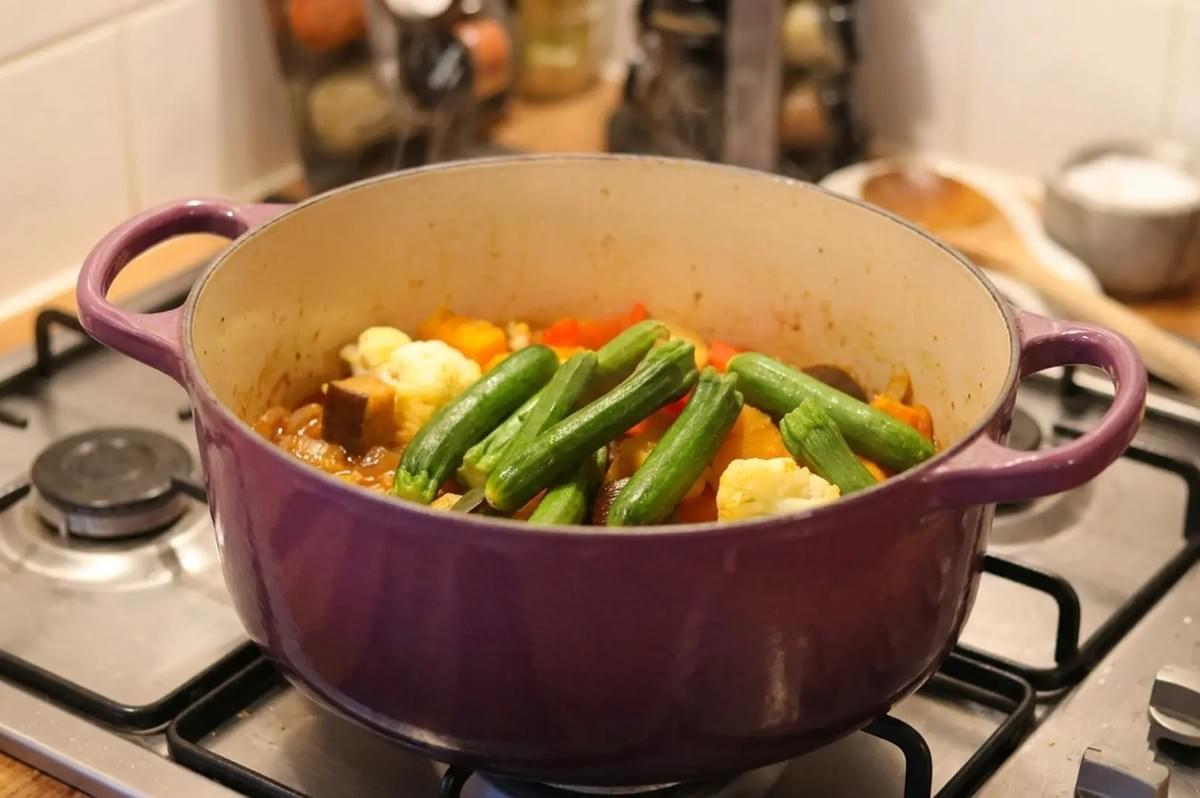 Purple Dutch oven on stovetop filled with simmering vegetables including green beans, cauliflower, and carrots Purple Dutch oven on stovetop filled with simmering vegetables including green beans, cauliflower, and carrots