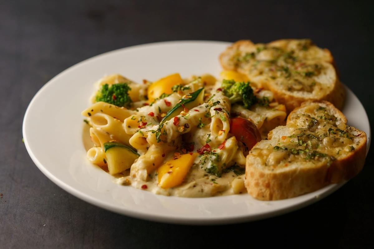 A white plate holds creamy penne pasta tossed with broccoli florets, yellow bell peppers, zucchini, and chili flakes. Beside the pasta are two toasted, seasoned slices of garlic bread against a dark slate background. A white plate holds creamy penne pasta tossed with broccoli florets, yellow bell peppers, zucchini, and chili flakes. Beside the pasta are two toasted, seasoned slices of garlic bread against a dark slate background.