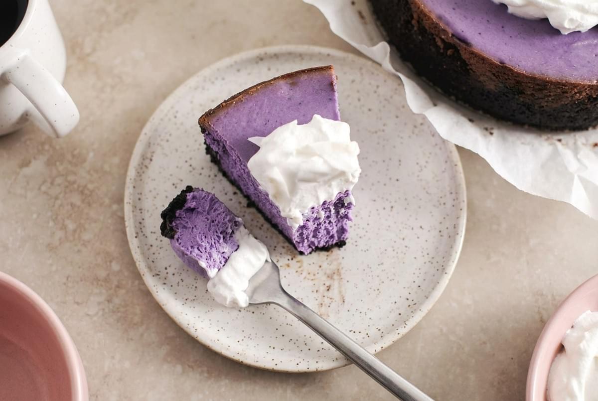 Slice of purple ube cheesecake topped with whipped cream, with a small bite taken on a fork. Slice of purple ube cheesecake topped with whipped cream, with a small bite taken on a fork.