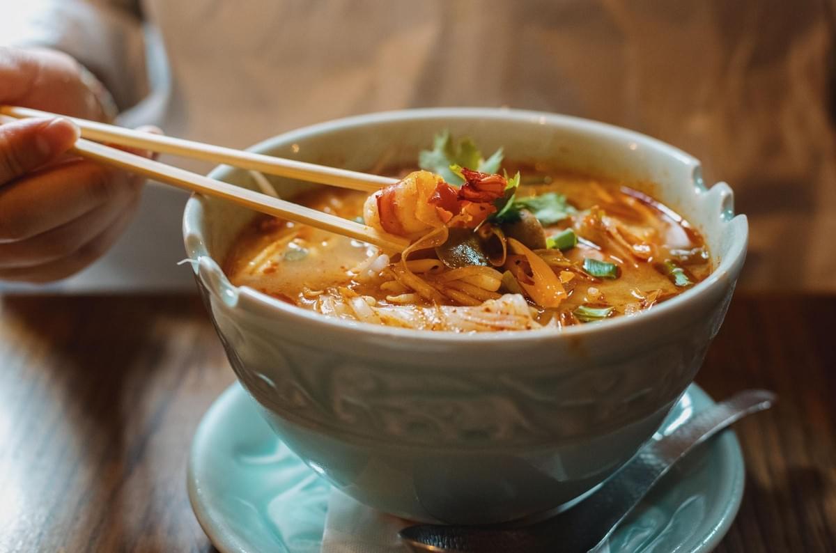 A hand uses wooden chopsticks to lift a cooked shrimp from a steaming bowl of noodle soup. The rich dish is served in a light green ceramic bowl resting on a matching saucer atop a wooden table. A hand uses wooden chopsticks to lift a cooked shrimp from a steaming bowl of noodle soup. The rich dish is served in a light green ceramic bowl resting on a matching saucer atop a wooden table.