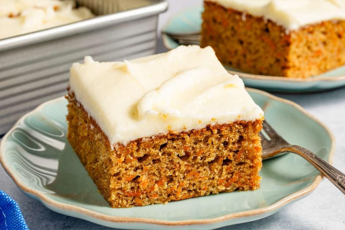 This image showcases a square slice of moist carrot cake topped with a thick, smooth layer of creamy white frosting. The cake is served on a light blue plate, with a metal baking pan visible in the background. This image showcases a square slice of moist carrot cake topped with a thick, smooth layer of creamy white frosting. The cake is served on a light blue plate, with a metal baking pan visible in the background.