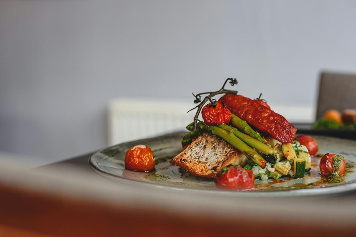A pan-seared fish fillet is elegantly plated alongside asparagus spears, roasted vine tomatoes, and a grilled sausage link. The dish is set on a dark ceramic plate drizzled with pesto and pan juices. A pan-seared fish fillet is elegantly plated alongside asparagus spears, roasted vine tomatoes, and a grilled sausage link. The dish is set on a dark ceramic plate drizzled with pesto and pan juices.