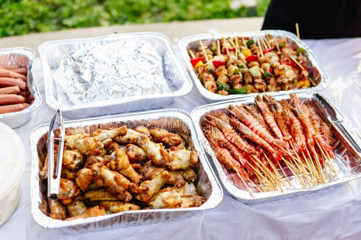 Various grilled meats and seafood, including chicken wings, shrimp skewers, and kebabs, are arranged in silver foil trays on a white table. Metal tongs rest in the trays, ready for serving guests at what appears to be an outdoor gathering or buffet. Various grilled meats and seafood, including chicken wings, shrimp skewers, and kebabs, are arranged in silver foil trays on a white table. Metal tongs rest in the trays, ready for serving guests at what appears to be an outdoor gathering or buffet.