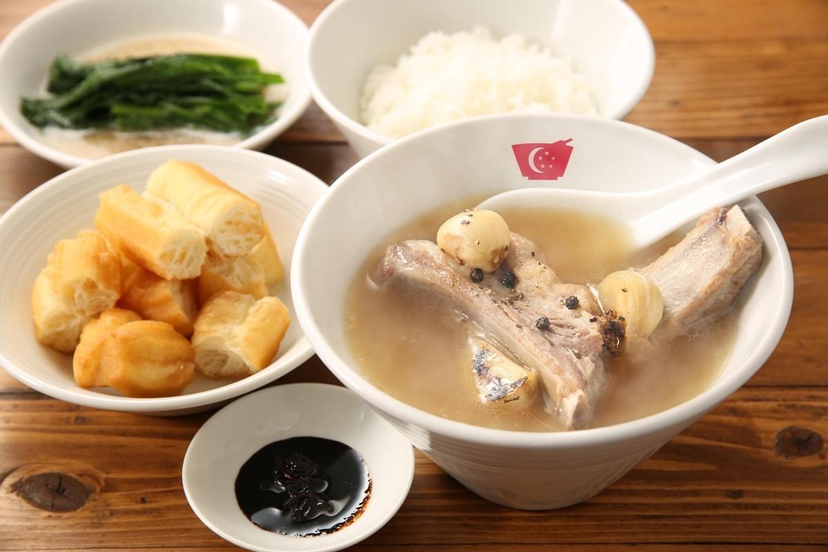 A large bowl of pork rib soup with garlic cloves and peppercorns sits on a wooden table with a white spoon inside. It is accompanied by side dishes including sliced fried dough fritters, leafy green vegetables, steamed white rice, and a small dish of dark sauce. A large bowl of pork rib soup with garlic cloves and peppercorns sits on a wooden table with a white spoon inside. It is accompanied by side dishes including sliced fried dough fritters, leafy green vegetables, steamed white rice, and a small dish of dark sauce.
