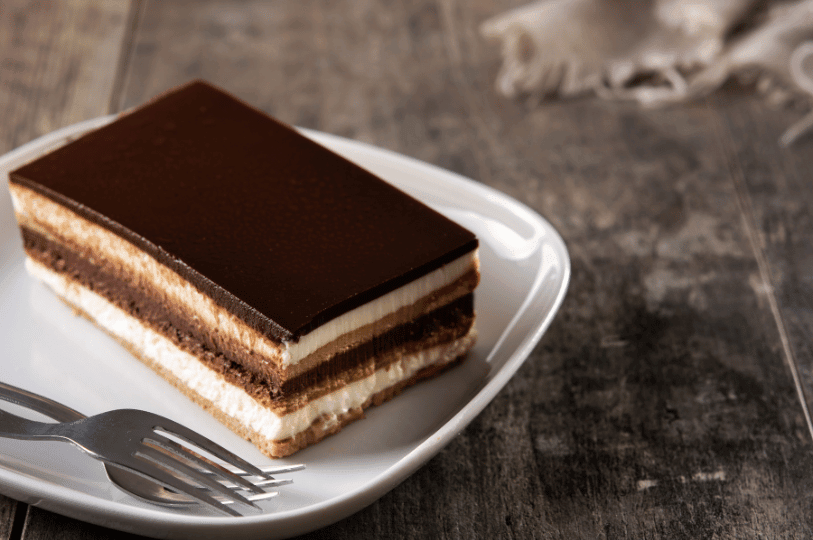 A close-up of a neat rectangular slice of opera cake on a square white plate, clearly showing its alternating layers of sponge, coffee buttercream, and chocolate ganache under a smooth chocolate glaze, with two forks resting beside it. A close-up of a neat rectangular slice of opera cake on a square white plate, clearly showing its alternating layers of sponge, coffee buttercream, and chocolate ganache under a smooth chocolate glaze, with two forks resting beside it.