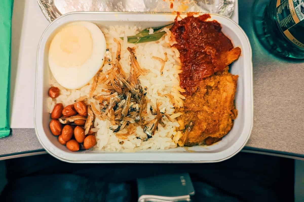 An overhead view shows an airplane meal tray containing Nasi Lemak, featuring a bed of rice topped with a hard-boiled egg slice, fried anchovies, peanuts, and a green leaf garnish. On the right side of the container, there is a portion of chicken curry alongside a vibrant red sambal sauce. An overhead view shows an airplane meal tray containing Nasi Lemak, featuring a bed of rice topped with a hard-boiled egg slice, fried anchovies, peanuts, and a green leaf garnish. On the right side of the container, there is a portion of chicken curry alongside a vibrant red sambal sauce.