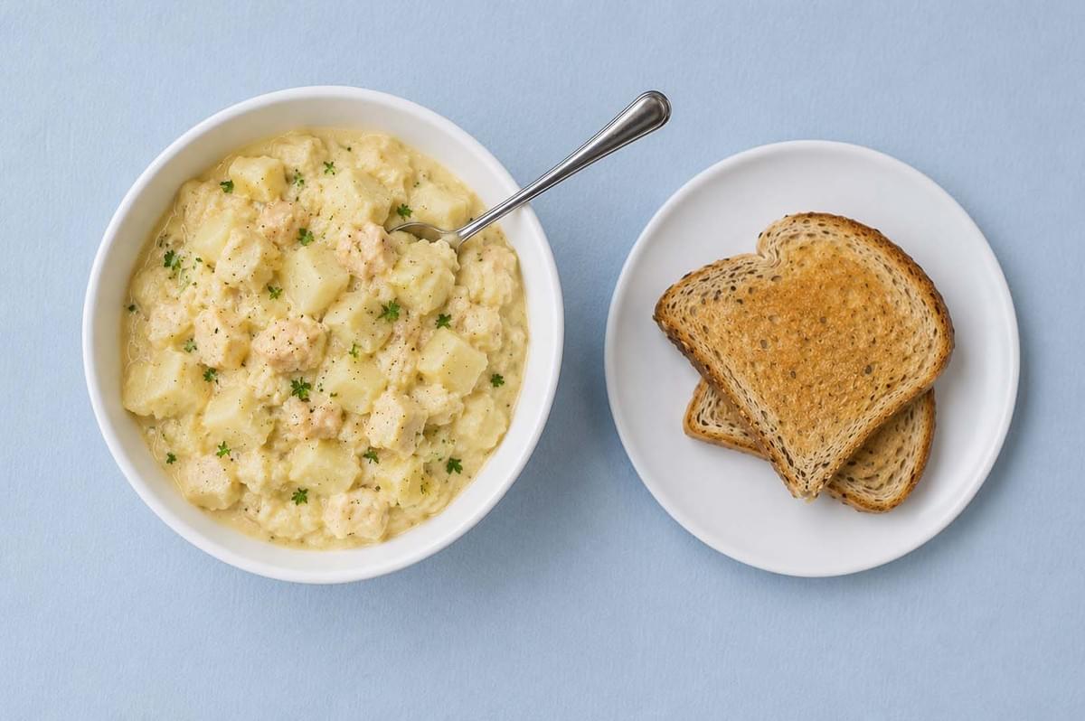 Top-down view of a white bowl filled with creamy fish and potato stew next to a plate of toasted bread Top-down view of a white bowl filled with creamy fish and potato stew next to a plate of toasted bread