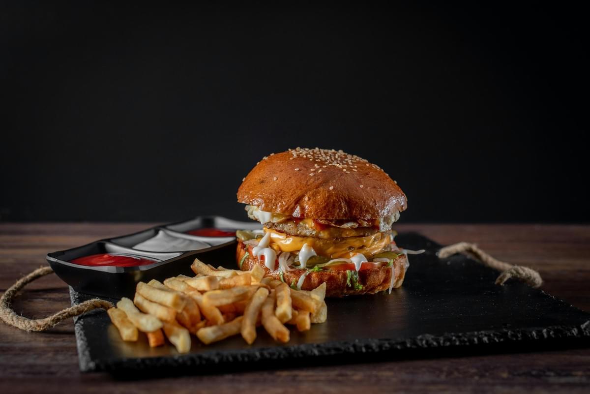 A juicy, fully stacked burger with melted cheese, lettuce, and sauce sits on a black slate tray next to a pile of golden French fries. Beside the meal is a small tray filled with dipping sauces, set against a dark wooden backdrop. A juicy, fully stacked burger with melted cheese, lettuce, and sauce sits on a black slate tray next to a pile of golden French fries. Beside the meal is a small tray filled with dipping sauces, set against a dark wooden backdrop.