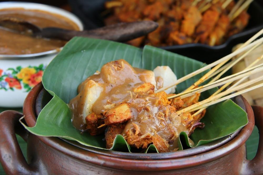 An up-close shot showcases chicken satay skewers and compressed rice cakes served on a vibrant green banana leaf inside a traditional clay bowl. Thick peanut sauce and crispy fried shallots cover the dish, with additional satay and a bowl of extra sauce visible in the blurred background. An up-close shot showcases chicken satay skewers and compressed rice cakes served on a vibrant green banana leaf inside a traditional clay bowl. Thick peanut sauce and crispy fried shallots cover the dish, with additional satay and a bowl of extra sauce visible in the blurred background.