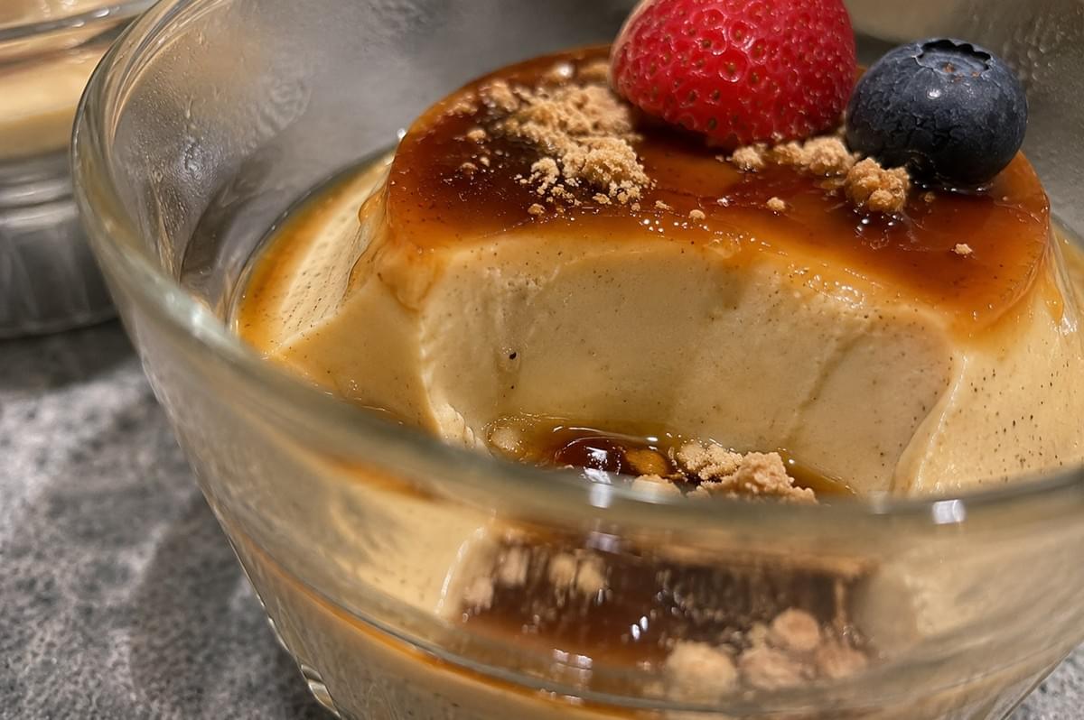 An extreme macro shot showing a scoop taken out of a smooth, creamy hojicha panna cotta inside a glass dish, revealing its silken custard texture pooling with dark syrup, kinako powder crumbs, a strawberry slice, and a fresh blueberry on top. An extreme macro shot showing a scoop taken out of a smooth, creamy hojicha panna cotta inside a glass dish, revealing its silken custard texture pooling with dark syrup, kinako powder crumbs, a strawberry slice, and a fresh blueberry on top.