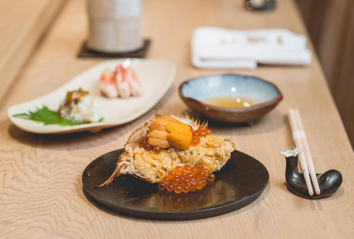 A crab shell filled with crab meat, seasoned rice, sea urchin, and bright orange salmon roe sits on a black plate atop a wooden table. In the background, chopsticks rest on a small holder alongside additional small sushi dishes and a dipping bowl. A crab shell filled with crab meat, seasoned rice, sea urchin, and bright orange salmon roe sits on a black plate atop a wooden table. In the background, chopsticks rest on a small holder alongside additional small sushi dishes and a dipping bowl.