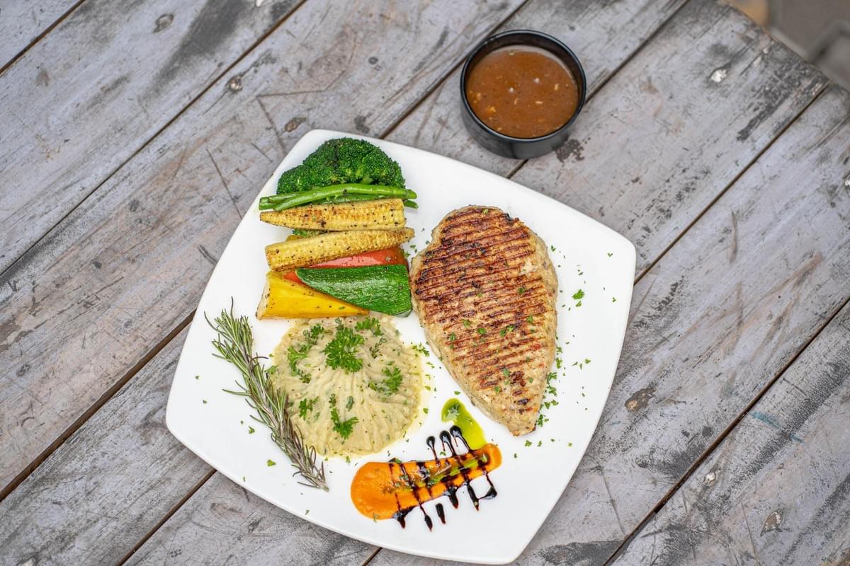 A square white plate on a weathered gray wooden table holds a grilled chicken breast with distinct grill marks alongside a side of mashed potatoes and an assortment of vegetables including broccoli, baby corn, green beans, and bell peppers. A small black bowl filled with brown gravy sits in the upper right corner of the frame. A square white plate on a weathered gray wooden table holds a grilled chicken breast with distinct grill marks alongside a side of mashed potatoes and an assortment of vegetables including broccoli, baby corn, green beans, and bell peppers. A small black bowl filled with brown gravy sits in the upper right corner of the frame.