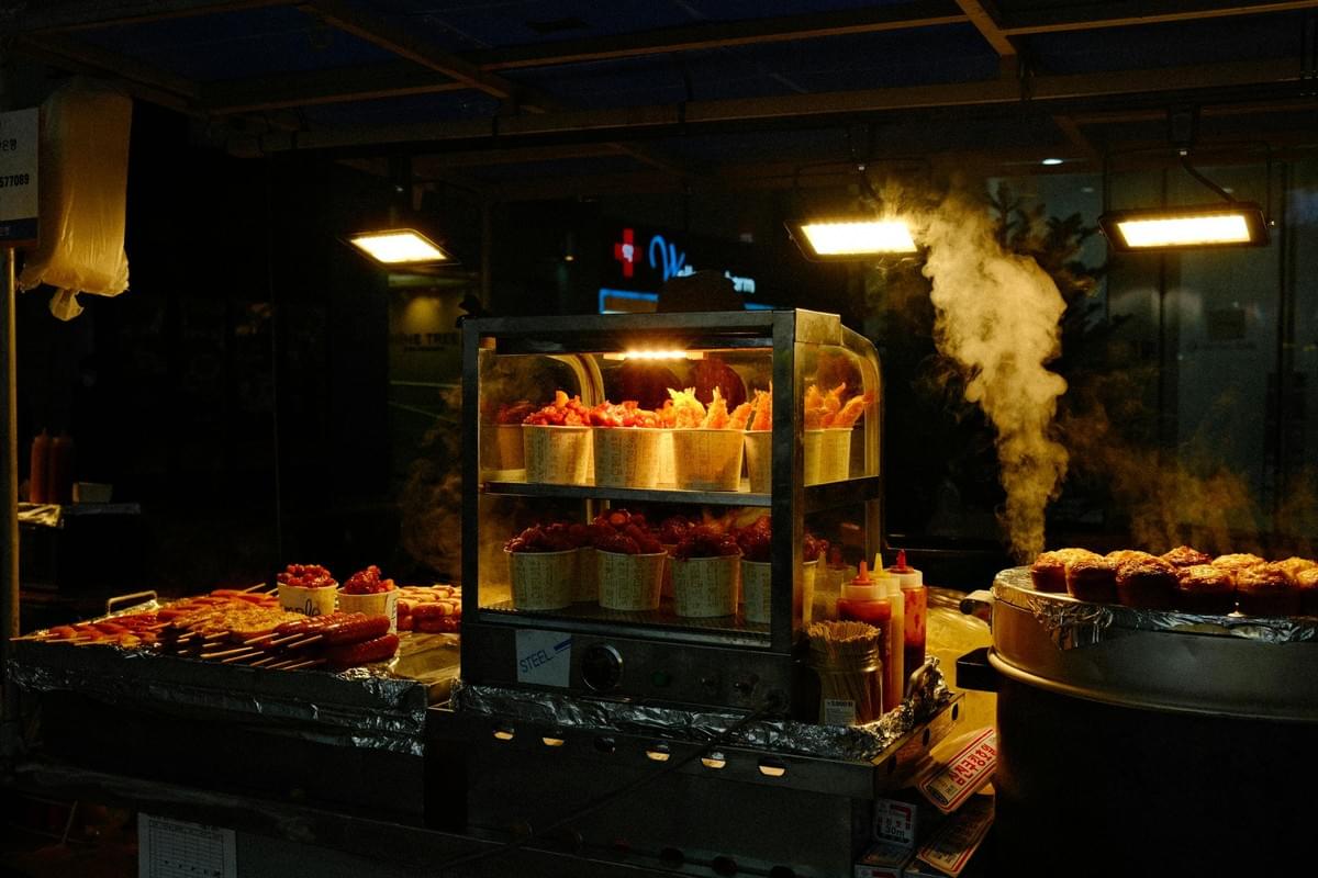 A street food stall at night glows under warm lights, showcasing cups of fried food inside a heated display case alongside skewers of sausages. To the right, thick steam rises from a large, covered metal steamer holding freshly cooked buns or dumplings. A street food stall at night glows under warm lights, showcasing cups of fried food inside a heated display case alongside skewers of sausages. To the right, thick steam rises from a large, covered metal steamer holding freshly cooked buns or dumplings.