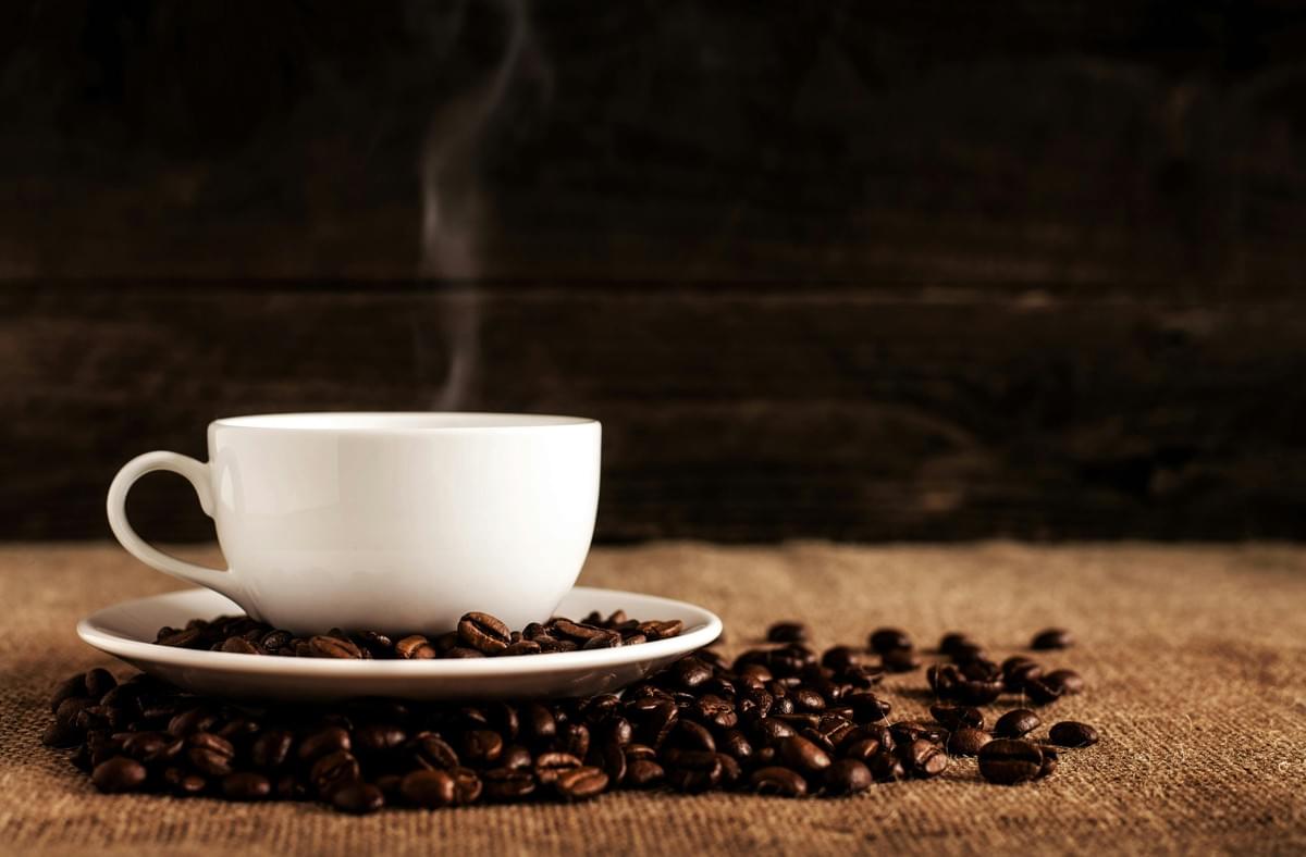 A steaming white ceramic cup sits on a saucer surrounded by scattered coffee beans. The scene is set on a textured burlap surface against a dark, rustic wooden background. A steaming white ceramic cup sits on a saucer surrounded by scattered coffee beans. The scene is set on a textured burlap surface against a dark, rustic wooden background.