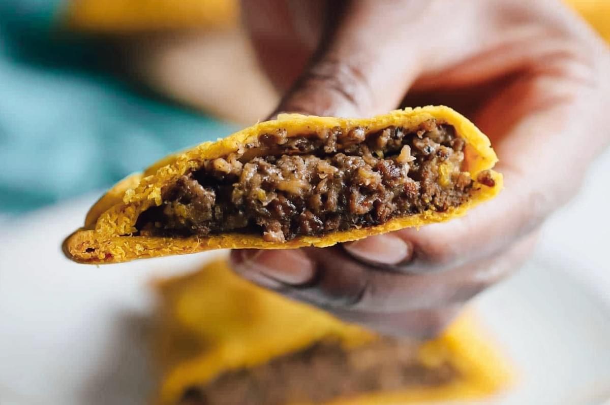 Eye-level close-up of a Jamaican beef patty held in hand, highlighting the curry-yellow pastry and rich, spicy ground beef filling. Eye-level close-up of a Jamaican beef patty held in hand, highlighting the curry-yellow pastry and rich, spicy ground beef filling.