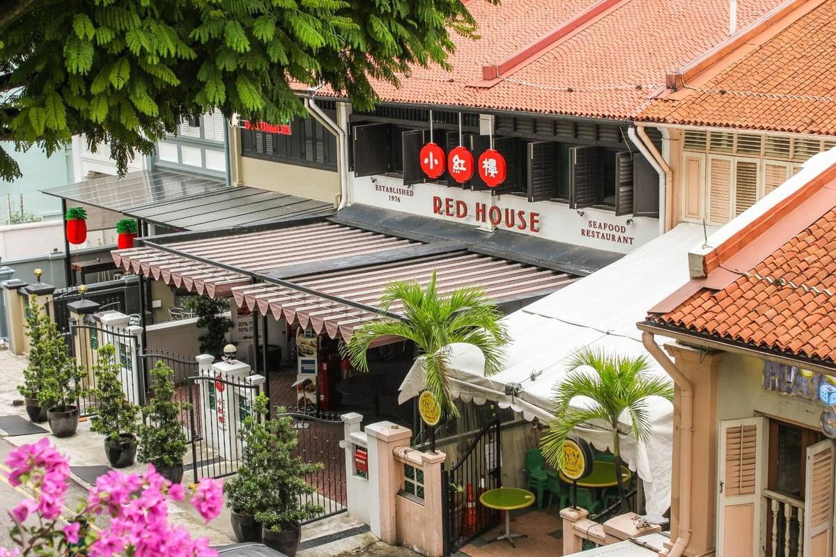 An elevated view of the Red House Seafood Restaurant featuring a traditional shophouse building with distinct orange tile roofing and black window shutters. The entrance is lined with small potted trees, bright pink bougainvillea flowers in the foreground, and palm trees under an awning. An elevated view of the Red House Seafood Restaurant featuring a traditional shophouse building with distinct orange tile roofing and black window shutters. The entrance is lined with small potted trees, bright pink bougainvillea flowers in the foreground, and palm trees under an awning.