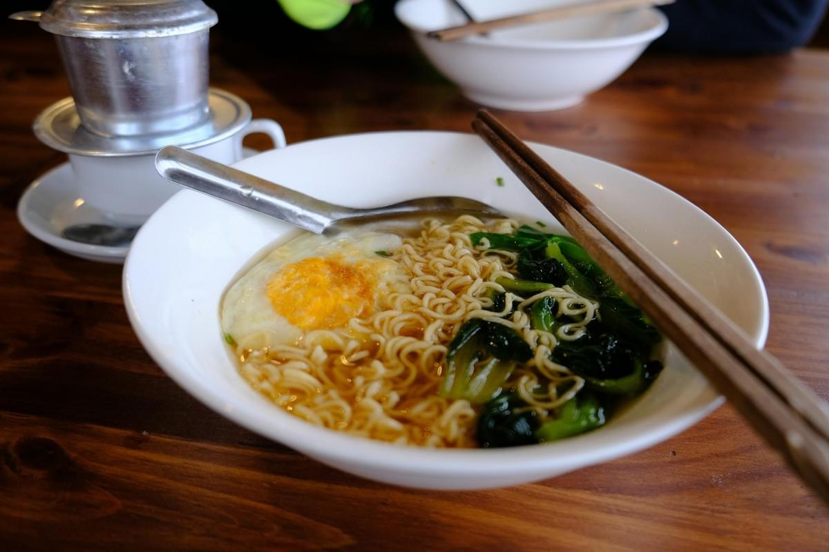 A white bowl filled with ramen noodle soup sits on a wooden table, topped with a fried egg and green leafy vegetables. A metal spoon and a pair of wooden chopsticks rest across the rim of the bowl, while a traditional Vietnamese coffee phin filter sits in the background. A white bowl filled with ramen noodle soup sits on a wooden table, topped with a fried egg and green leafy vegetables. A metal spoon and a pair of wooden chopsticks rest across the rim of the bowl, while a traditional Vietnamese coffee phin filter sits in the background.
