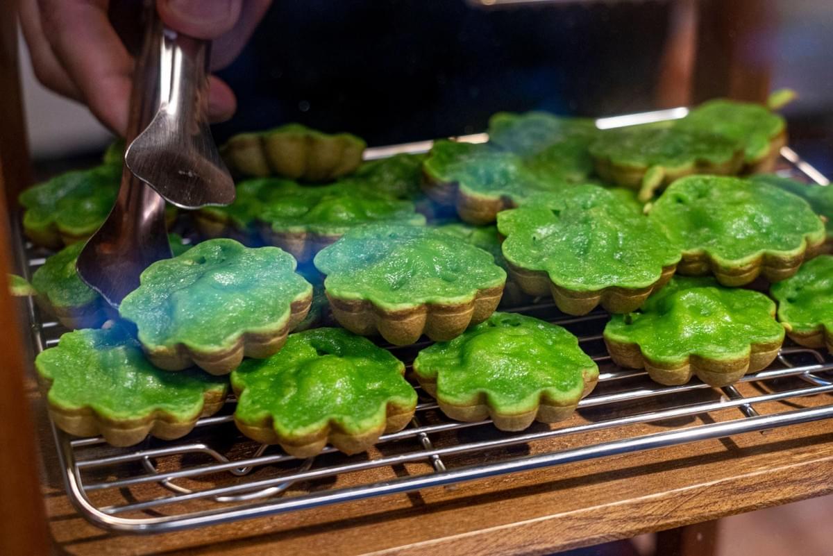 A hand uses metal tongs to pick up a flower-shaped, bright green pandan cake resting on a metal cooling rack. The rack is filled with numerous freshly baked treats, showing off their soft golden edges and vibrant green tops. A hand uses metal tongs to pick up a flower-shaped, bright green pandan cake resting on a metal cooling rack. The rack is filled with numerous freshly baked treats, showing off their soft golden edges and vibrant green tops.