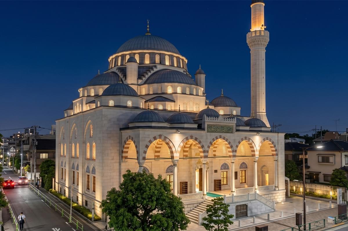 Exterior view of a beautiful, illuminated mosque with multiple domes and a tall minaret at twilight in a residential neighborhood. Exterior view of a beautiful, illuminated mosque with multiple domes and a tall minaret at twilight in a residential neighborhood.