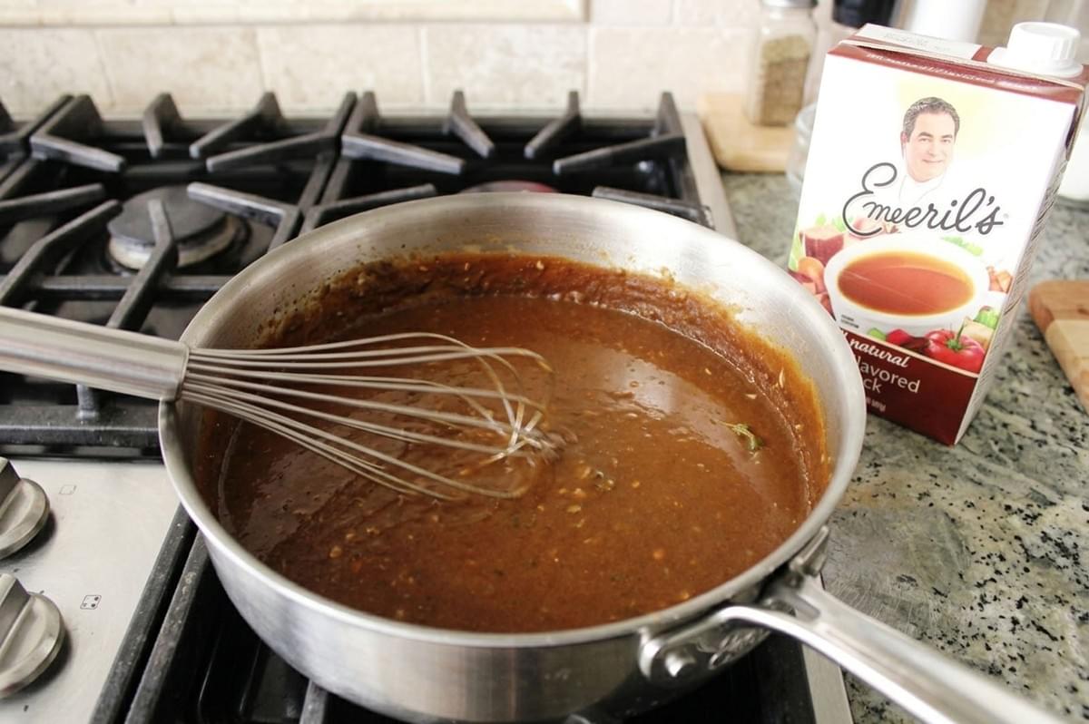 Dark brown roux being whisked in stainless steel pan on stovetop with stock carton nearby Dark brown roux being whisked in stainless steel pan on stovetop with stock carton nearby