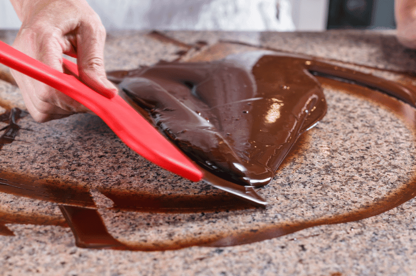 A pastry chef using a bright red spatula to work and spread glossy melted chocolate across a granite countertop for traditional table tempering. A pastry chef using a bright red spatula to work and spread glossy melted chocolate across a granite countertop for traditional table tempering.
