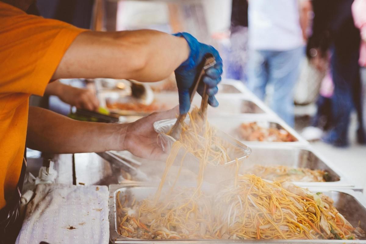 A person wearing a blue glove uses metal tongs to serve steaming noodles into a foil container from a large metal tray. The scene captures a busy food stall with several other trays of food visible in the blurred background. A person wearing a blue glove uses metal tongs to serve steaming noodles into a foil container from a large metal tray. The scene captures a busy food stall with several other trays of food visible in the blurred background.