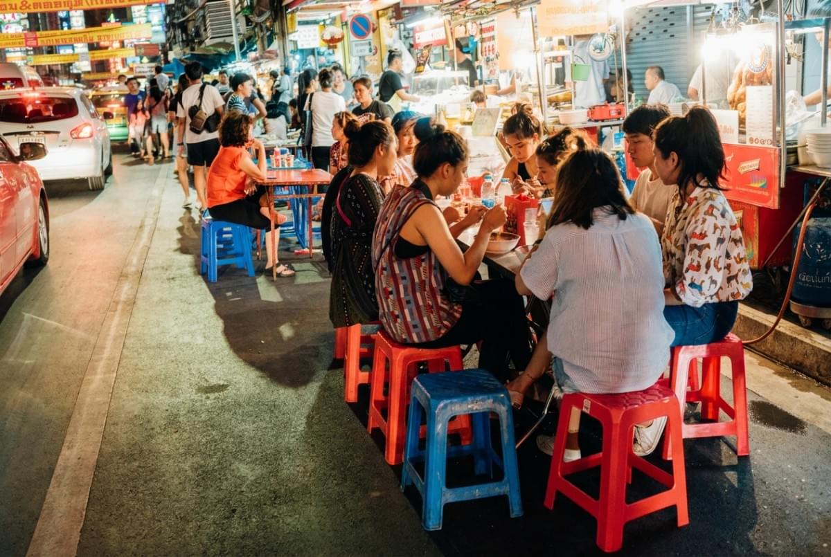 Diners sitting on colorful plastic stools at sidewalk tables along a busy street market at night. Diners sitting on colorful plastic stools at sidewalk tables along a busy street market at night.
