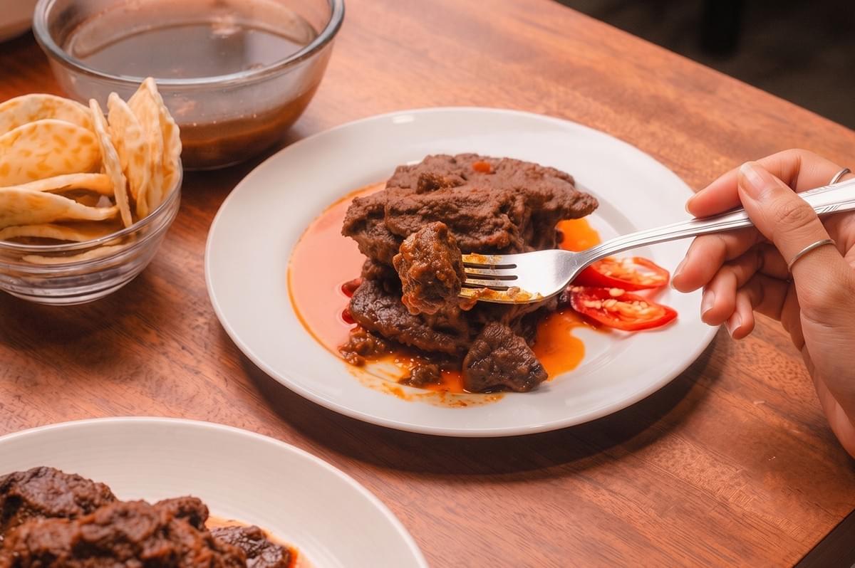 Person eating homemade beef rendang with a fork from a white plate. Person eating homemade beef rendang with a fork from a white plate.