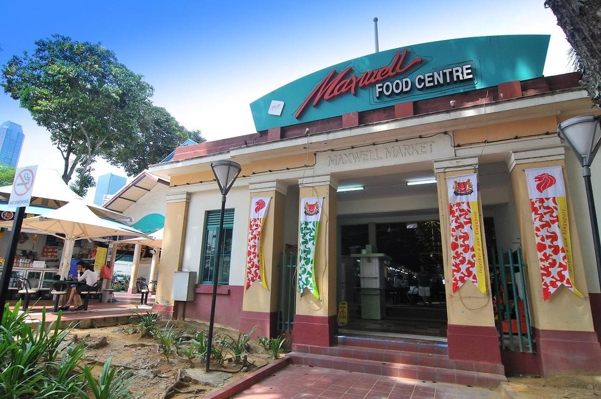 The exterior entrance of Maxwell Food Centre displays a prominent turquoise sign above the building's facade. Decorative banners hang along the front pillars, and outdoor seating with umbrellas sits adjacent to the walkway on a sunny day. The exterior entrance of Maxwell Food Centre displays a prominent turquoise sign above the building's facade. Decorative banners hang along the front pillars, and outdoor seating with umbrellas sits adjacent to the walkway on a sunny day.