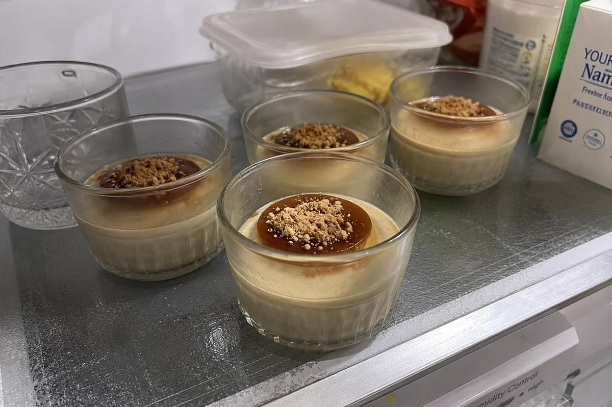 Four clear glass ramekins containing set hojicha panna cotta desserts—each garnished with dark kuromitsu syrup and fine kinako powder—chilling on a glass shelf inside a refrigerator. Four clear glass ramekins containing set hojicha panna cotta desserts—each garnished with dark kuromitsu syrup and fine kinako powder—chilling on a glass shelf inside a refrigerator.