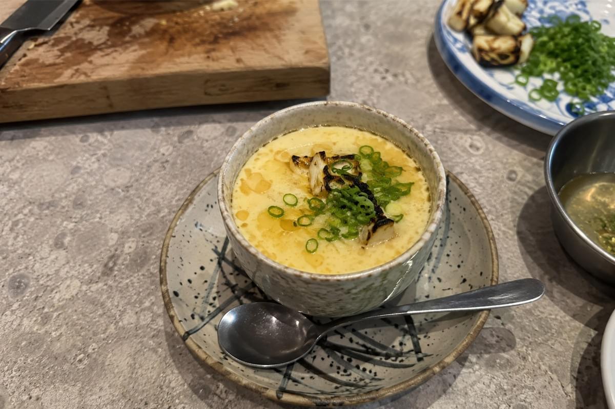 A top-down, high-angle shot shows a bowl of steamed egg custard (chawanmushi) served in a speckled cream-colored ceramic cup sitting atop a matching dish with abstract dark brushstrokes. The smooth, golden custard surface is glistening with droplets of ginger oil and garnished with pieces of charred leek and finely sliced green scallions. A silver spoon rests on the right side of the saucer, while the surrounding grey countertop features prep ingredients, including a wooden cutting board and extra chopped scallions on a blue-and-white plate. A top-down, high-angle shot shows a bowl of steamed egg custard (chawanmushi) served in a speckled cream-colored ceramic cup sitting atop a matching dish with abstract dark brushstrokes. The smooth, golden custard surface is glistening with droplets of ginger oil and garnished with pieces of charred leek and finely sliced green scallions. A silver spoon rests on the right side of the saucer, while the surrounding grey countertop features prep ingredients, including a wooden cutting board and extra chopped scallions on a blue-and-white plate.