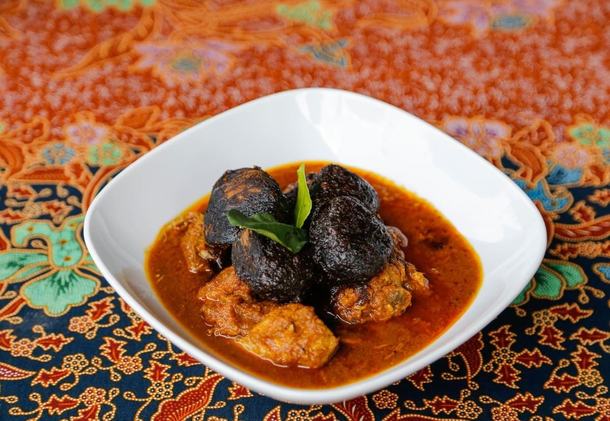 A white ceramic bowl contains a rich, orange-red curry dish topped with dark, whole buah keluak nuts and garnished with fresh green leaves. The bowl rests on a vibrant batik tablecloth featuring intricate orange, blue, and floral patterns. A white ceramic bowl contains a rich, orange-red curry dish topped with dark, whole buah keluak nuts and garnished with fresh green leaves. The bowl rests on a vibrant batik tablecloth featuring intricate orange, blue, and floral patterns.
