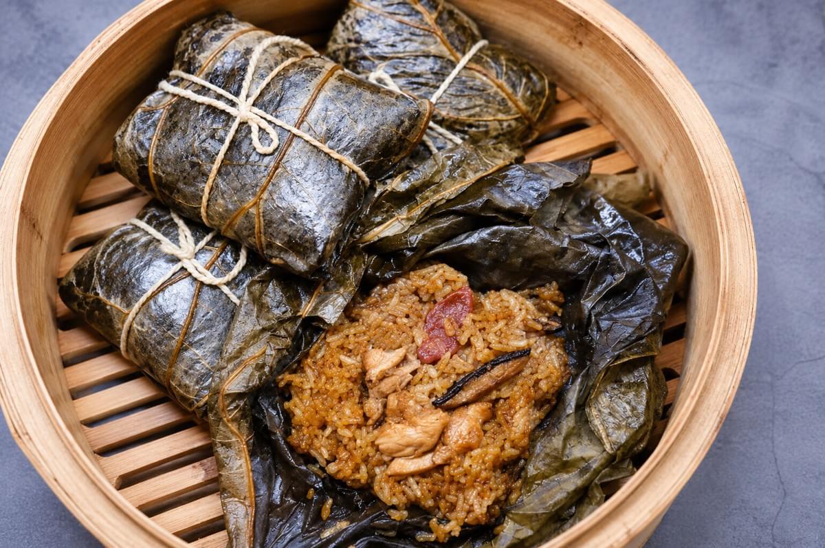 A top-down view of a round bamboo steamer containing three tied lotus leaf parcels and one unwrapped leaf revealing seasoned sticky rice with chicken, Chinese sausage, and mushrooms. A top-down view of a round bamboo steamer containing three tied lotus leaf parcels and one unwrapped leaf revealing seasoned sticky rice with chicken, Chinese sausage, and mushrooms.
