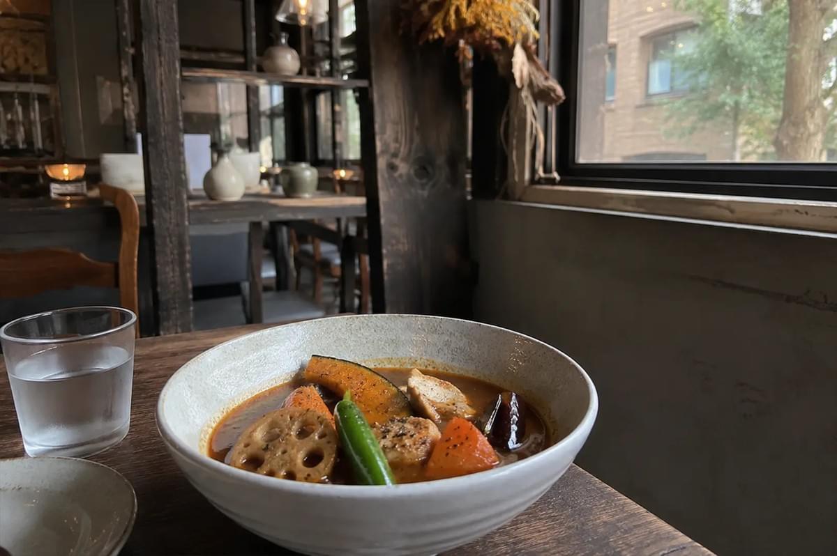 A bowl of steaming soup curry is the focal point, featuring colorful chunks of roasted kabocha squash, lotus root, a green chili, and tender pieces of meat submerged in a rich, reddish-brown broth. A clear glass of water sits to the left of the bowl on a dark wooden table, while the background reveals a rustic, atmospheric dining space with dark wood paneling, decorative ceramic vessels, a dried flower arrangement hanging near the window, and a view of trees outside. A bowl of steaming soup curry is the focal point, featuring colorful chunks of roasted kabocha squash, lotus root, a green chili, and tender pieces of meat submerged in a rich, reddish-brown broth. A clear glass of water sits to the left of the bowl on a dark wooden table, while the background reveals a rustic, atmospheric dining space with dark wood paneling, decorative ceramic vessels, a dried flower arrangement hanging near the window, and a view of trees outside.