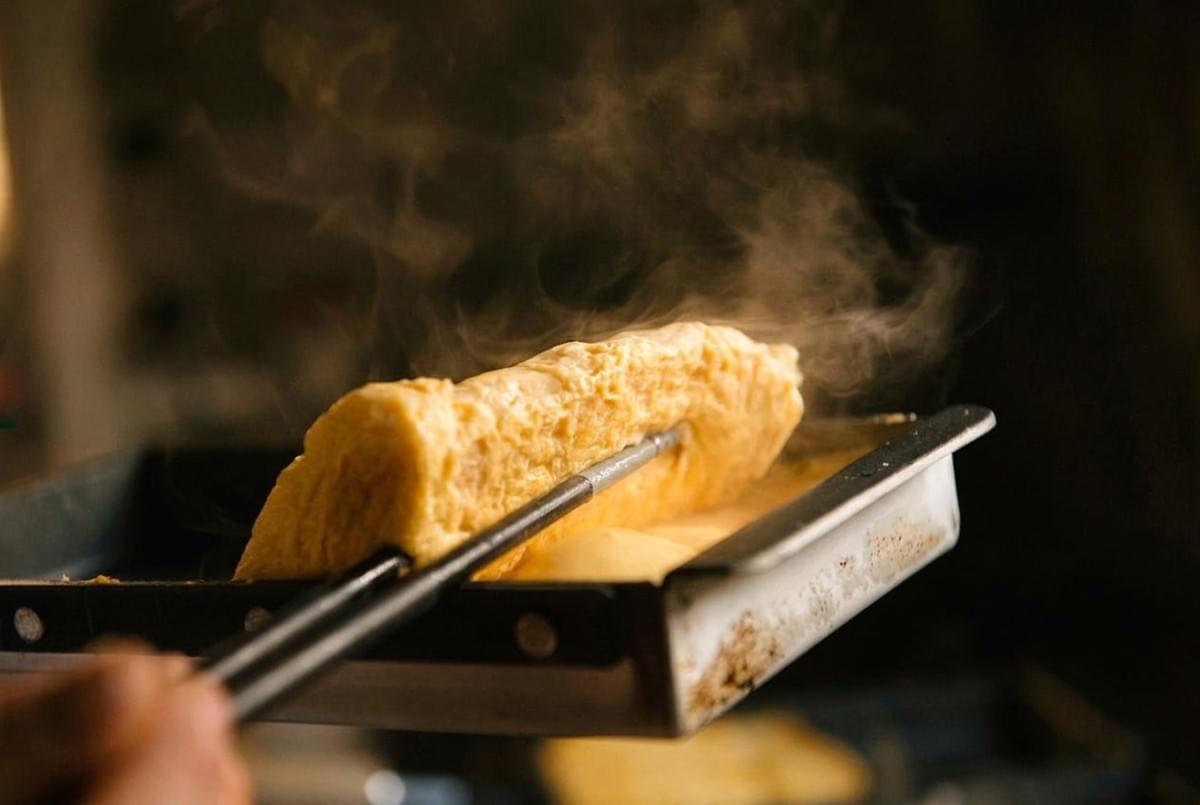 Steam rising as a hot rolled tamagoyaki omelet is lifted with chopsticks from a pan. Steam rising as a hot rolled tamagoyaki omelet is lifted with chopsticks from a pan.