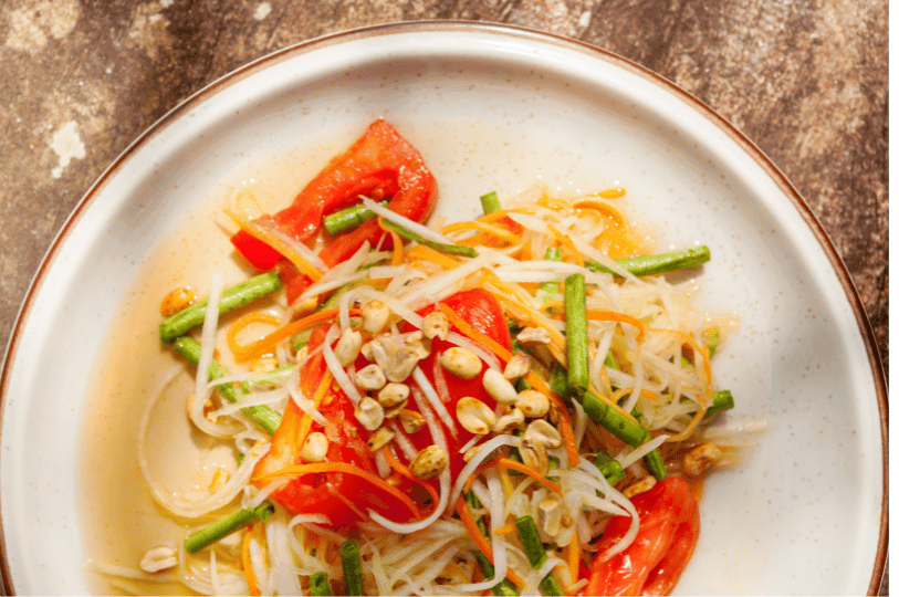 A top-down view of Som Tam (Papaya Salad) in a white bowl, showing shredded green papaya mixed with bright red tomato wedges, long beans, and roasted peanuts in a light dressing. A top-down view of Som Tam (Papaya Salad) in a white bowl, showing shredded green papaya mixed with bright red tomato wedges, long beans, and roasted peanuts in a light dressing.
