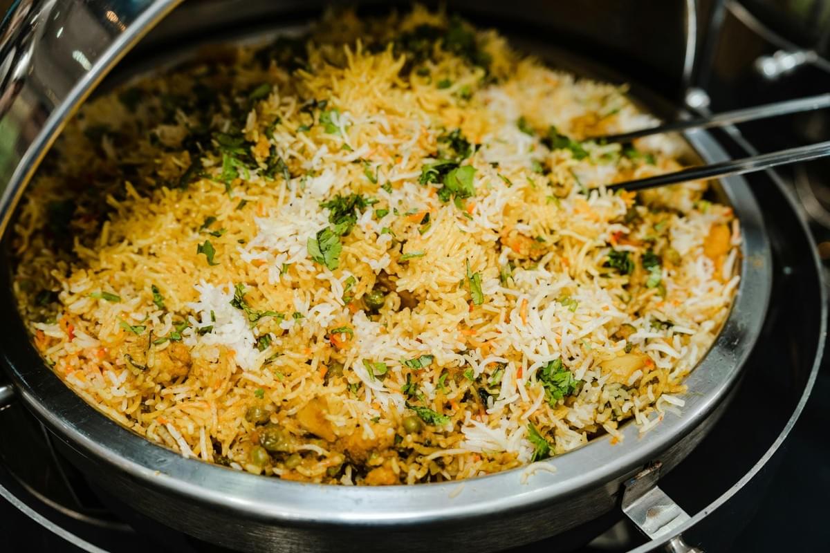 A large silver chafing dish is filled with fragrant biryani rice topped with fresh cilantro. Metal serving utensils rest on the rim, ready for serving at a buffet setup. A large silver chafing dish is filled with fragrant biryani rice topped with fresh cilantro. Metal serving utensils rest on the rim, ready for serving at a buffet setup.