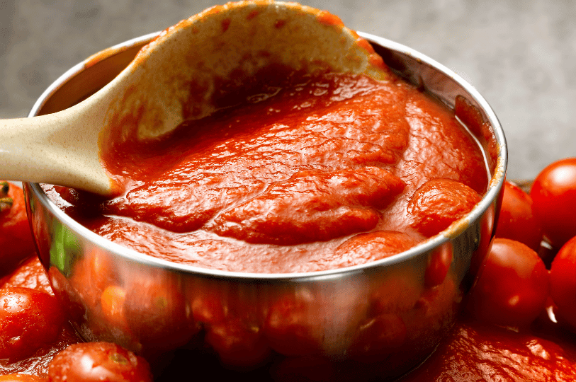 A close-up of a stainless steel bowl filled with a chunky, vibrant red Sauce Tomate, with fresh cherry tomatoes scattered around the base and a large spoon scooping the sauce. A close-up of a stainless steel bowl filled with a chunky, vibrant red Sauce Tomate, with fresh cherry tomatoes scattered around the base and a large spoon scooping the sauce.