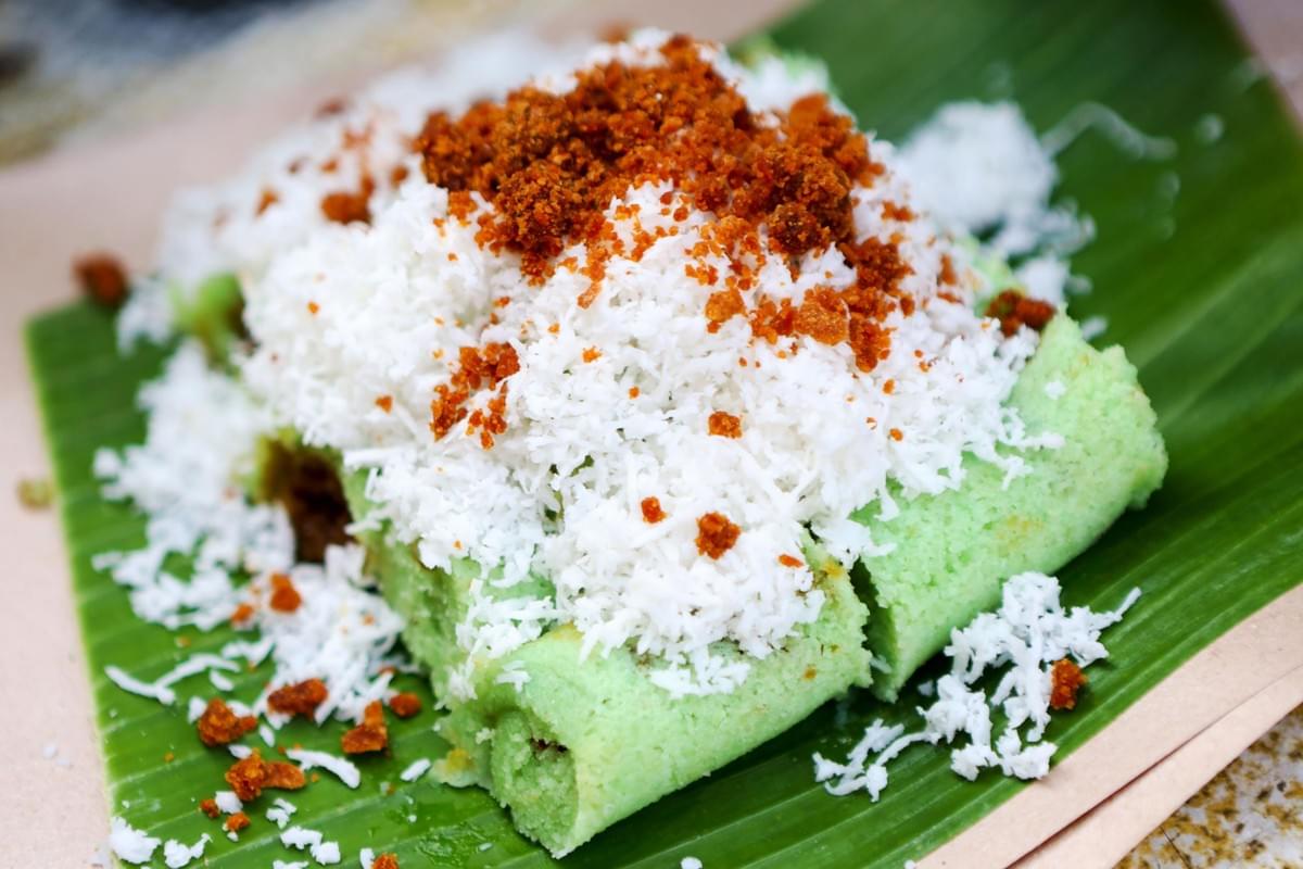 A close-up shot shows light green rice cakes served on a fresh banana leaf. They are generously topped with freshly grated white coconut and sprinkled with brown palm sugar crumbs. A close-up shot shows light green rice cakes served on a fresh banana leaf. They are generously topped with freshly grated white coconut and sprinkled with brown palm sugar crumbs.